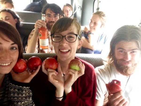 Image of several students in van seats, including Claire Jordy, who sits at the center and has light brown hair and glasses. The students in the front row smile and hold apples up by their faces. One boy in the middle row holds up an orange beverage in a class container. 