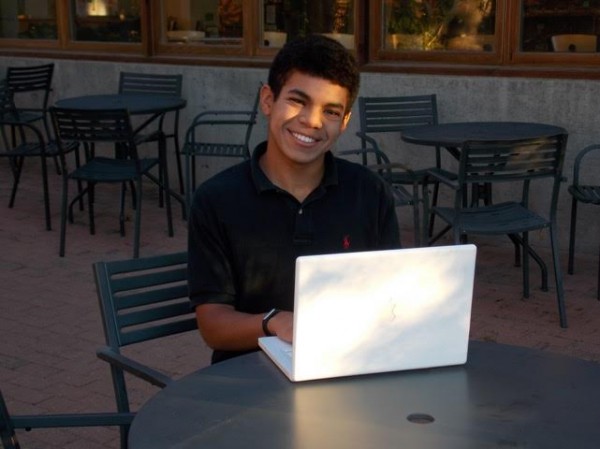 Image of Emmanuel Greeno, a boy with light brown skin and dark hair, smiling and sitting at a cafe table typing on an Apple laptop. 