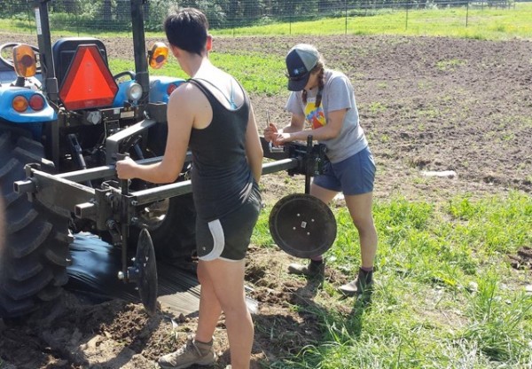 Image of two students repairing a tractor outside a plowed field. The student to the left has short-cropped hair and wears a black tank top and shorts. The student to the right has braids and wears a tan-and-blue baseball cap, grey T-shirt, blue shorts, and boots. 