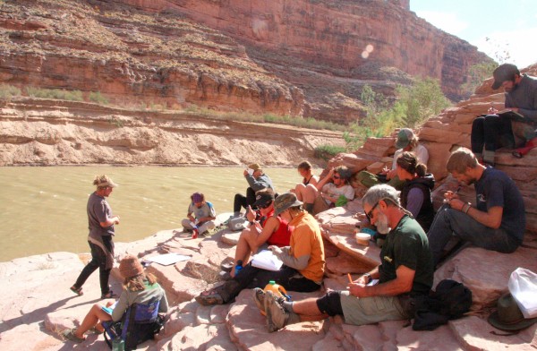 Image of many students sitting at different levels on a hill of clay-colored rocks next to a brown river. They are participating in an outdoor adventure for the Wilderness Exploration Landscape course.