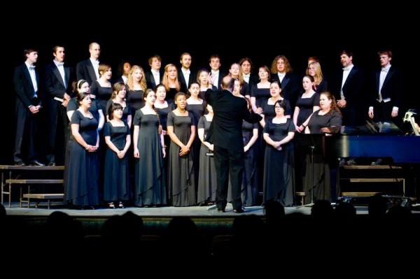 Image of a choir of Green Mountain College students, wearing blue and black dresses and tuxedoes, arranged on a multi-leveled riser and singing. A conductor stands in front of them with his hands raised. 
