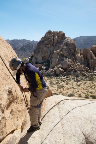 Image of a male student rigging his rappel with desert shrubbery and large rock mountains in the background. The student wears a blue long-sleeved shirt, khakis, and a grey helmet, and looks away from the camera to focus on the rappel.