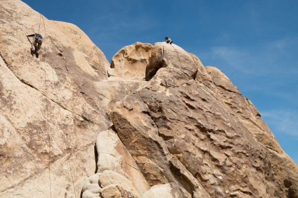Image of Andew and another student scaling a rock face in Joshua Tree National Park as part of their outdoor adventure experience. One student climbs on a rope, while the other rests at the top of a rock. The two students can barely be identified on the massive rocks.