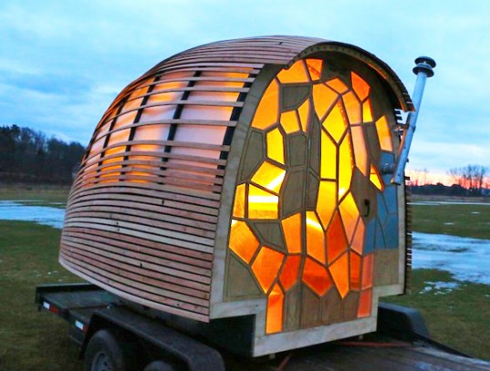 Image of a wheeled "tiny house" with an exterior made of warm-colored stained glass. The house sits on a trailer bed in a grassy field with some remains of snow at sunset.