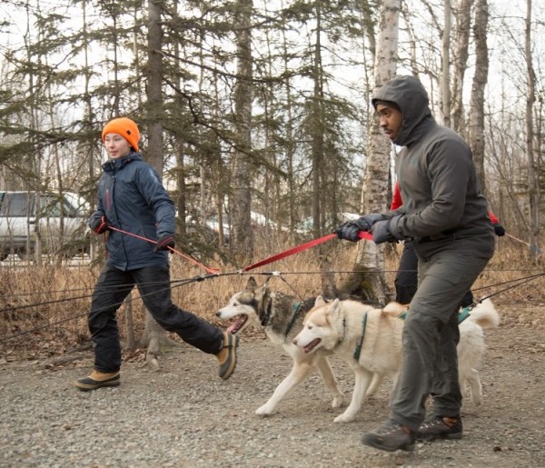 Image of two students learning to mush husky dogs on a forest trail. The student to the left has pale skin, an orange hat, blue coat, and black pants. The student to the right has dark skin, wears a hooded grey jacket, and long grey pants.