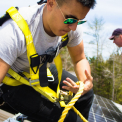 Image of a male student from College of the Atlantic installing solar panels. He has short brown hair and wears sunglasses, a grey T-shirt, and a yellow safety harness. He clutches a yellow rope in one hand. 