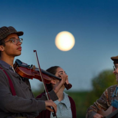 Image of Dickinson students Max Jacobs, Gaby Corcoran, and Alexander Dillan standing in front of a full moon on the farm. Max is playing a violin. Gaby and Alexander are laughing at each other as if in the middle of a joke as Alexander plays the guitar. 