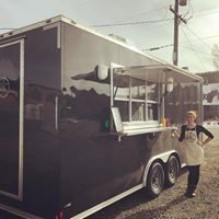 Image of Kara Kessler, a white woman with light brown hair, standing outside a black vegan food truck on a dirt road. She wears a black long sleeve shirt and pants and a white apron, and puts her right hand on her hip. 