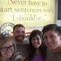 Image of Travis Sherlin standing with two female and another male student, all of whom are smiling. They are standing in front of a Peace Corps sign that says "Never have to start sentences with 'I should've'...". 