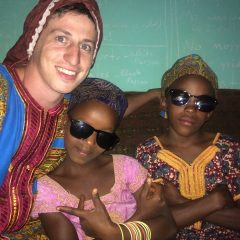 Image of Travis Sherlin, a white boy with short-cropped brown hair, sitting with two dark-skinned girls with sunglasses. The three are wearing colorful robes and smiling and posing for the camera. Travis wears a red head covering, and the two girls next to him wear brown rounded caps.