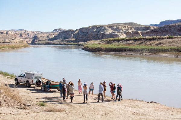 Image of a group of about a dozen young people standing in a circle next to a rowboat hooked to a white truck on a dusty road. In the background are a long river, a grassy bank, and several rock formations. 