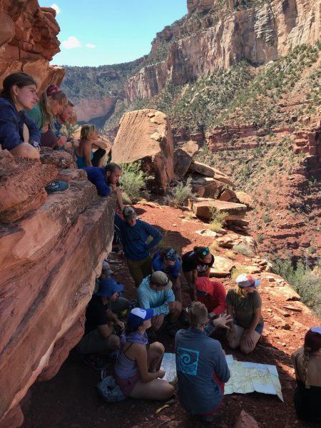 Image of about a dozen students sitting across a vertical reddish rock formation which makes up the left third of the image. The students all appear to be looking to the right. In the background, greens sprout on a multi tiered red canyon face. 