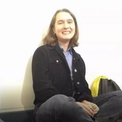Image of Leigh Rankin, a young white woman with short brown hair, a black jacket, blue shirt, and grey pants, sitting cross-legged up against a white wall and laughing.